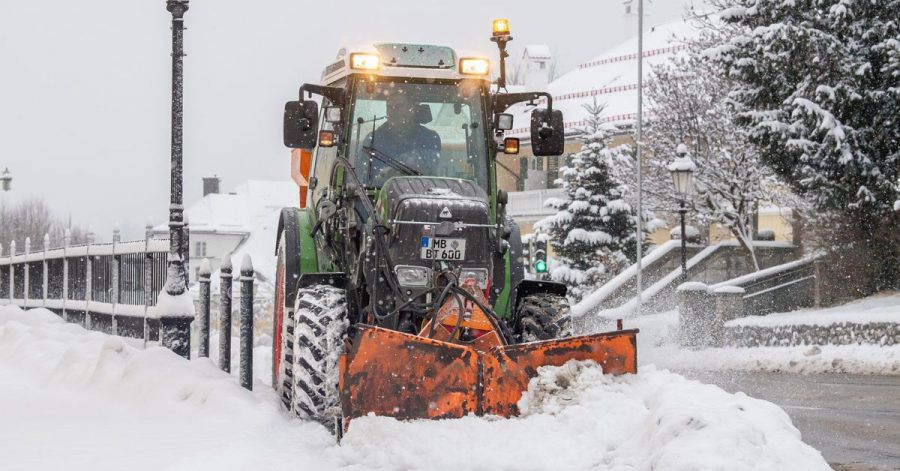 Ein Traktor mit Schneepflug räumt eine verschneite Straße im Ortskern der Ortschaft am Tegernsee.