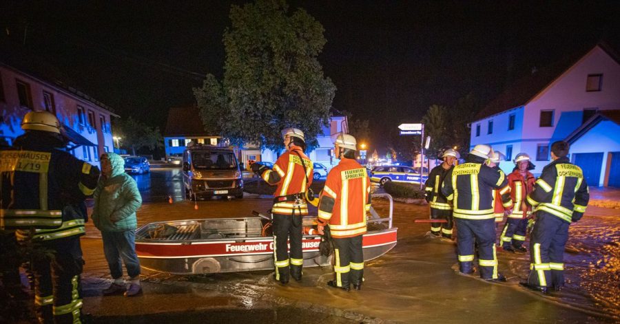 Feuerwehrleute im Kreis Biberach stehen neben einem Feuerwehrboot auf einer Straße in den Fluten eines heftigen Unwetters.