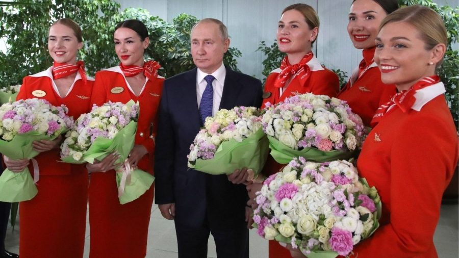 Wladimir Putin umringt von Stewardessen in roter Uniform mit Blumensträußen